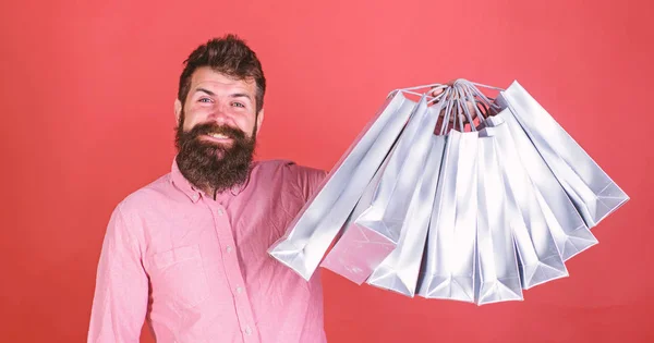 Mann mit Bart und Schnurrbart hält Einkaufstaschen, roter Hintergrund. Hipster mit glücklichem Gesicht sind shoppingsüchtig oder shopaholic. Typen, die in der Verkaufssaison mit Rabatten einkaufen. Einkaufskonzept — Stockfoto