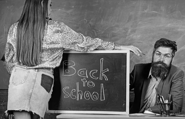 Δάσκαλος αυστηρή κάθονται τραπέζι chalkboard φόντο. Μαθητής σε μίνι φούστα με ωραίο πισινό σταθεί κοντά μαυροπίνακα. Σχολική πειθαρχία και κανόνες συμπεριφοράς. Μαθήτρια ξελογιάζει έμπειρη δασκάλα — Φωτογραφία Αρχείου