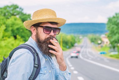 Eski alışkanlık. Sakal ve bıyık hasır şapka Sigara İçilmeyen, defocused yol arka plan içinde olan adam. Gezgin şık hipster fren ile sigara al. Uzun bir yolculuk önce Sigara İçilmeyen