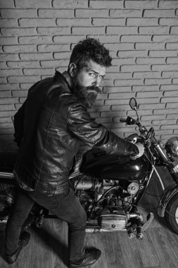 Yenilikçi, vahşi Motorcu deri ceketli sıkı yüzündeki motosiklet üzerinde oturur. Sakallı, motorcu motorlu bisiklet garaj, tuğla duvar arka plan yakınındaki deri ceketli adam. Erkeksi hobi kavramı