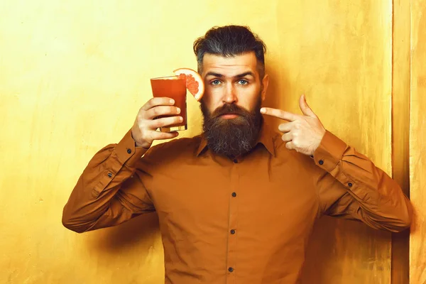 Bearded man, long beard. Brutal caucasian serious hipster with moustache in brown shirt holding tropical alcoholic fresh cocktail with orange piece on golden texture background