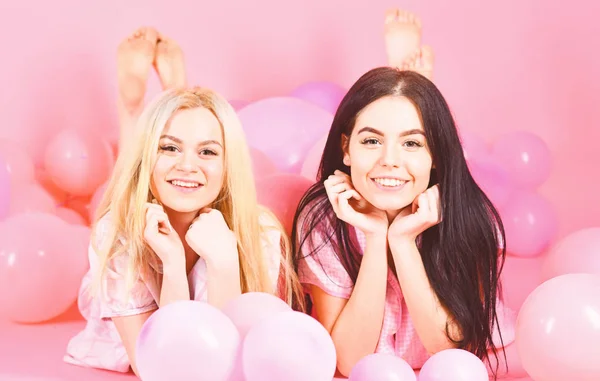 Κορίτσια Βάλτε στην κοιλιά κοντά σε μπαλόνια, ροζ φόντο. Έννοια του κουτσομπολιού. Ξανθιά και μελαχρινή σε χαμογελαστά πρόσωπα να διασκεδάσουν στο κόμμα εγχώριων κρεβατοκάμαρων. Αδελφές ή καλύτεροι φίλοι στο πιτζάμες στο κοριτσίστικο Πιτζάμες κόμμα — Φωτογραφία Αρχείου