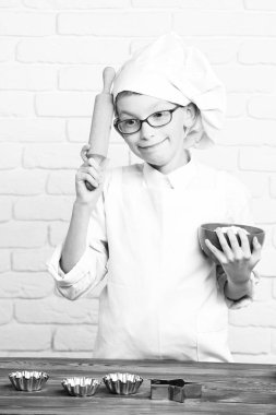 Genç çocuk küçük sevimli aşçı Şef beyaz üniforma ve Şapka kalıpları tabloyla yakınındaki kek için ayakta ve oklava tuğla duvar adam üzerinde kırmızı bir kase tutarak gözlüklü komik surat