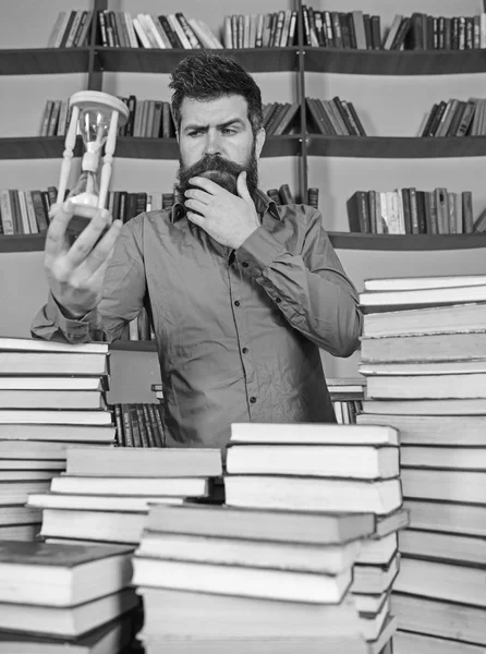 Conceito de bibliotecário. Homem no rosto atencioso fica entre pilhas de livros, enquanto mantém ampulheta, estantes de livros no fundo. Professor, cientista com barba está à mesa com livros, desfocado — Fotografia de Stock