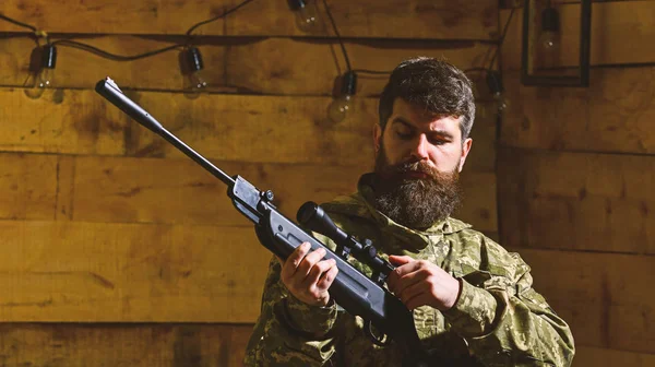 Huntsman concept. Macho on strict face at gamekeepers house. Man with beard wears camouflage clothing, wooden interior background. Hunter, brutal hipster with gun prepare rifle for hunting, copy spac — Stock Photo, Image
