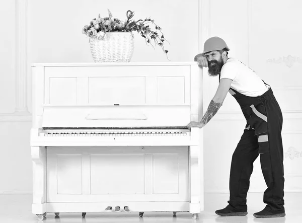 Hombre con barba trabajador en casco y overoles empuja, esfuerzos para mover el piano, fondo blanco. El cargador mueve el instrumento de piano. Concepto de cargas pesadas. Courier entrega muebles, se muda, se traslada —  Fotos de Stock