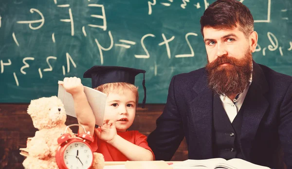 Far med skägg, lärare lär son, kid boy. Undervisning barn koncept. Lärare och elev i mortarboard, svarta tavlan på bakgrund. Kid studier med lärare, nära häfte, klocka, Leksak — Stockfoto