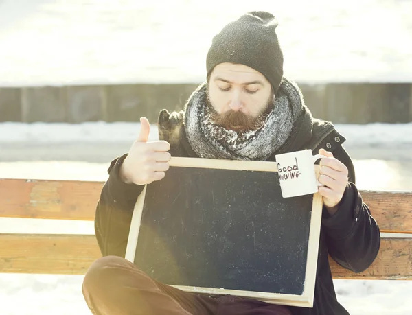 Stilig förvånad man, hipster med skägg och mustasch täckt med vit frost, håller tom svarta tavlan och kopp med god morgon text vinterdag på naturlig bakgrund, kopiera spac — Stockfoto