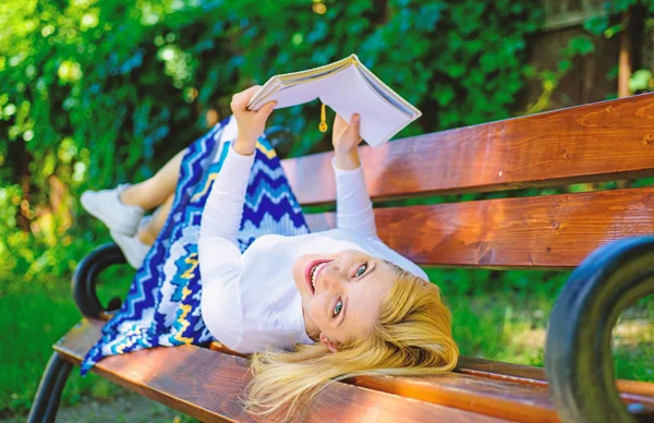 Interesting book. Lady smiling face enjoy rest. Girl reading outdoors while relaxing on bench. Girl lay bench park relaxing with book, green nature background. Woman spend leisure with book