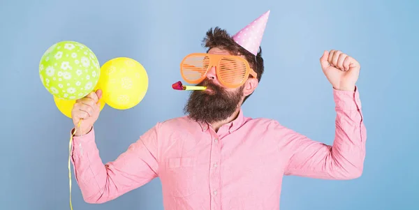 Bearded man in birthday cap and huge crazy glasses dancing, party hard, fun time concept. Hipster with trimmed beard and mustache posing with party whistle and bright balloons on blue background