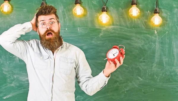 Esqueci-me do conceito do tempo. Homem com barba e bigode na expressão facial confusa na sala de aula. O professor de óculos tem despertador. Barbudo hipster detém relógio, quadro no fundo — Fotografia de Stock