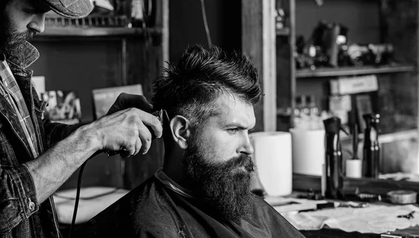 Hipster client getting haircut. Haircut concept. Barber styling hair of bearded client with comb and clipper. Barber with hair clipper works on hairstyle for man with beard, barbershop background — Stock Photo, Image
