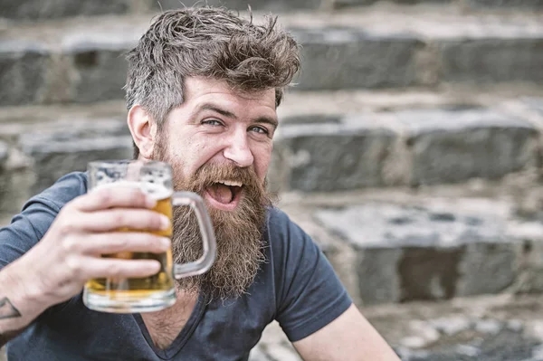 Hipster with long beard looks relaxed. Bearded hipster holds beer mug, drinks beer outdoor. Man with beard and mustache on happy face, stony background, defocused. Craft beer concept