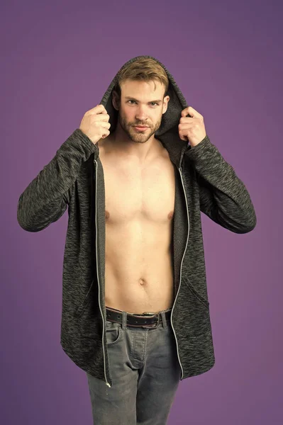 Hombre en la cara sonriente intenta chaqueta con capucha, fondo violeta. Chico con cerdas en la cara y la piel lisa en el pecho se prueba la ropa. Concepto de moda. El hombre se ve atractivo en chaqueta larga con capucha — Foto de Stock