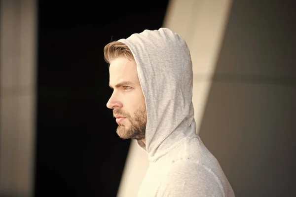 Guy bearded and attractive hooded, side view Man with bristle serious face, urban background, defocused. Needs to stay alone. Man beard unshaven guy looks handsome hooded. Walk alone concept