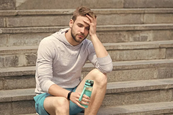Working out on stairs. Man athletic appearance exhausted holds water bottle. Man athlete sport clothes refreshing. Athlete drink water training at stadium sunny day. Exhausting workout concept