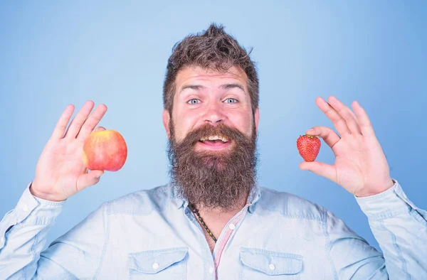 Beneficios de verano. Hombre barbudo sonriente sostiene manzana y fresa en las manos fondo azul. La fruta y la baya en las manos la alternativa sana. Concepto nutricional de la vitamina fruta. Vitamina para dietas sanitarias — Foto de Stock