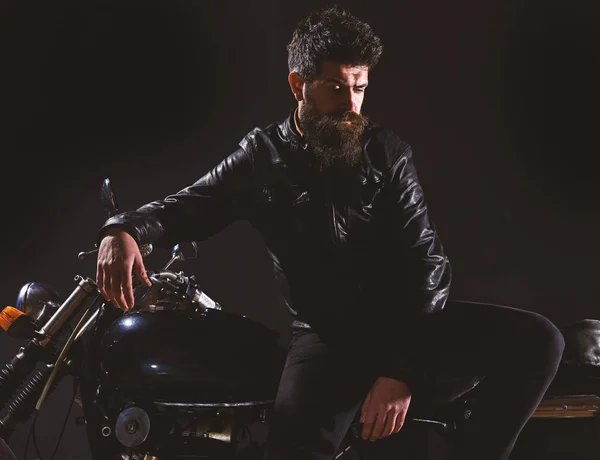 Macho, brutal biker in leather jacket stand near motorcycle at night time. Brutality and masculine concept. Man with beard, biker in leather jacket lean on motor bike in darkness, black background