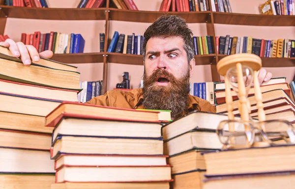 Professor ou estudante com barba senta-se à mesa com livros, desfocado. Conceito bibliófilo. Homem no rosto confuso entre pilhas de livros, enquanto estudava na biblioteca, estantes de livros no fundo — Fotografia de Stock