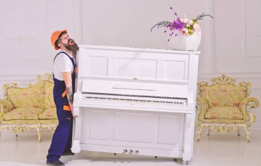 Courier delivers furniture, move out, relocation. Heavy loads concept. Man with beard worker in helmet and overalls lifts up, efforts to move piano, white background. Loader moves piano instrument clipart