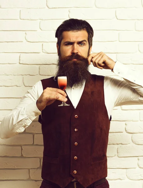 handsome bearded man with long beard and mustache has stylish hair on serious face holding glass of alcoholic shot in vintage suede leather waistcoat on white brick wall studio background