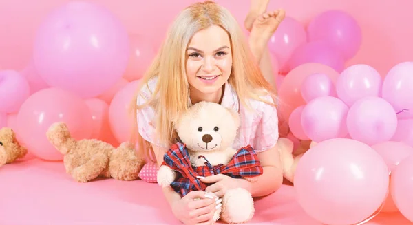 Blonde on smiling face relaxing with teddy bear toy. Woman cute celebrate birthday with balloons. Girl in pajama, domestic clothes lay near air balloons, pink background. Birthday girl concept