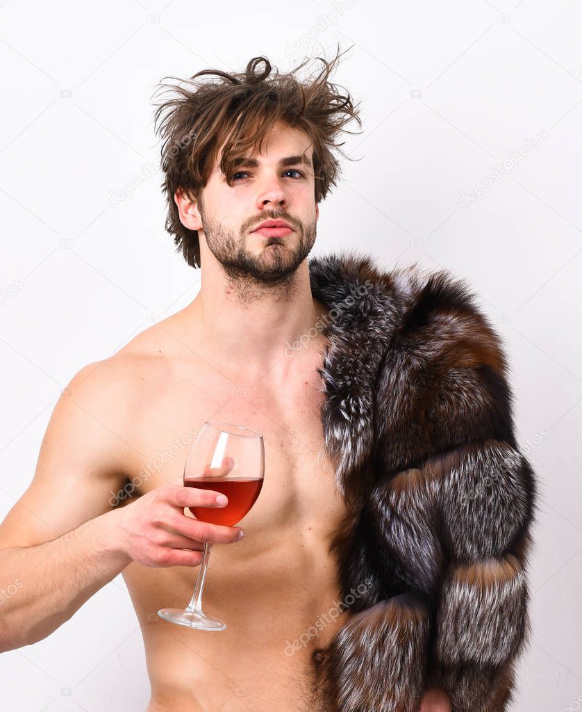 Sexy sleepy macho tousled hair drink wine or alcohol isolated on white. Luxury status symbol. Luxury lifestyle and wellbeing. Richness and luxury concept. Guy attractive posing fur coat on naked body