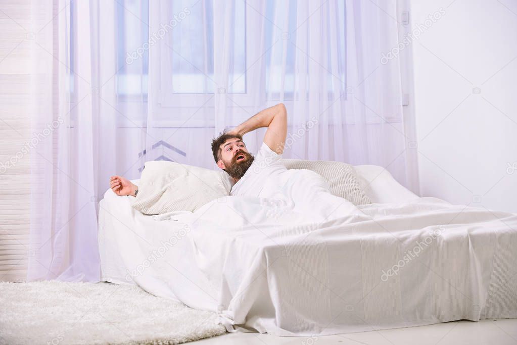 Man in shirt laying on bed awake, white curtain on background. Guy on shocked face waking up in morning. Macho with beard and mustache overslept waking up call. Wake up and oversleep concept