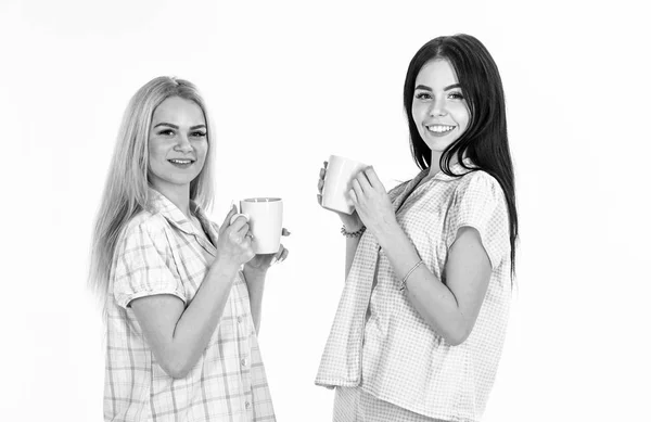 Sisters or best friends in pajamas. Morning coffee concept. Blonde and brunette on smiling faces holds mugs with coffee. Girls drinking tea or coffee in morning, isolated on white background — Stock Photo, Image