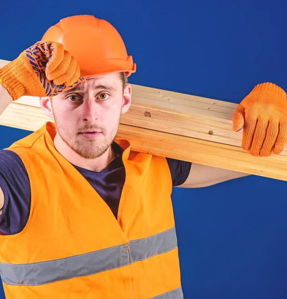 Concept de menuisier. Menuisier, menuisier, ouvrier, constructeur porte une poutre en bois sur l'épaule. Homme en gants de protection tient la visière du casque, corrige le casque dur sur la tête, fond bleu, gros plan — Photo