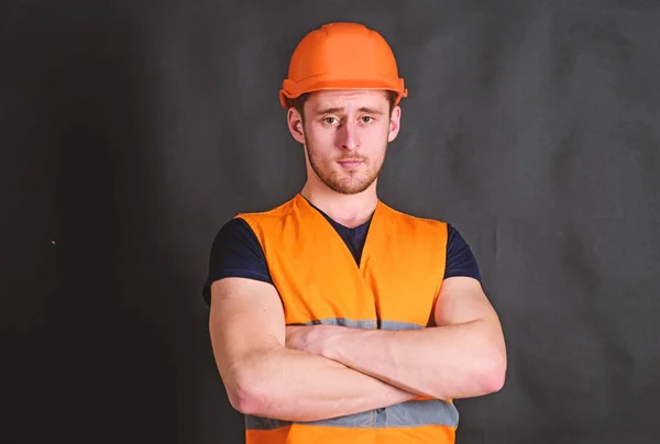 Man in helmet, hard hat hold arms crossed on chest, black background. Worker, contractor, builder on strict face with muscular biceps. Strong builder concept. Builder in helmet posing, copy space