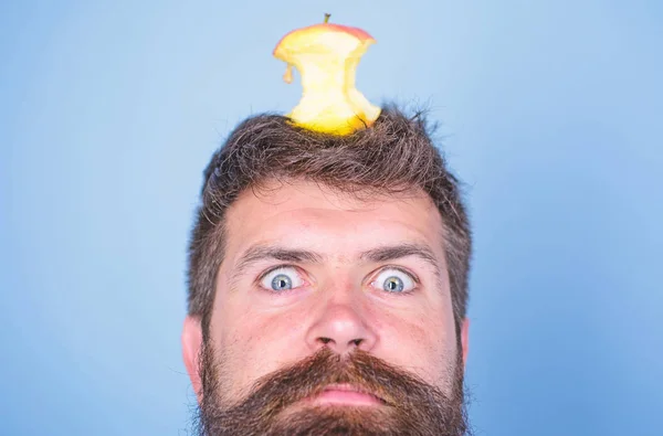 Hipster shocked face with apple stump target on head blue background, close up. Man handsome hipster long beard almost eaten apple stump on head as target. Weight loss goal. Live target concept