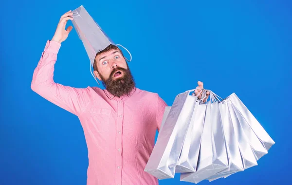 Hombre con barba y bigote lleva bolsas de compras, fondo azul. Hombre de compras en temporada de ventas con descuentos. Concepto de compras. Hipster en la cara sorprendida con bolsa en la cabeza es adicto a las compras —  Fotos de Stock