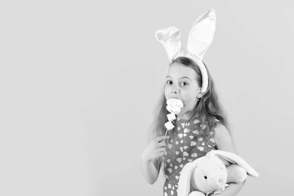 Easter and spring concept. Child with rabbit in bunny ears headband. Holiday gifts and presents. Childhood, youth and growth. Girl eating lollipop candy on pink background, copy spac — Stock Photo, Image