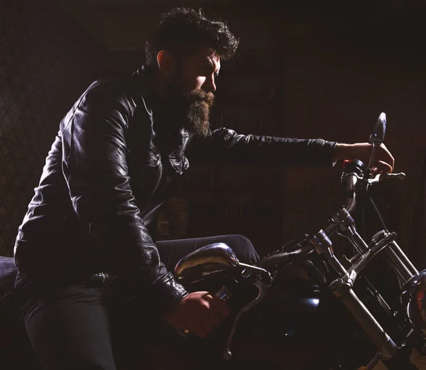 Macho, brutal biker in leather jacket riding motorcycle at night time, copy space. Man with beard, biker in leather jacket sitting on motor bike in darkness, black background. Night racer concept
