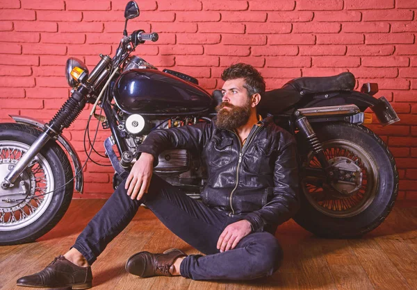 Man with beard, biker in leather jacket near motor bike in garage, brick wall background. Hipster, brutal biker on pensive face in leather jacket sit on floor near motorcycle. Brutal biker concept