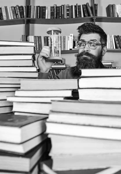 Man on surprised face holds hourglass while studying, bookshelves on background. Time flow concept. Teacher or student with beard studying in library. Man, scientist in glasses looks at hourglass — Stock Photo, Image