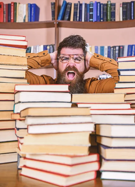 Concept de découverte scientifique. Enseignant ou élève barbu porte des lunettes, s'assoit à table avec des livres, déconcentré. Homme au visage choqué entre des piles de livres à la bibliothèque, étagères sur fond — Photo