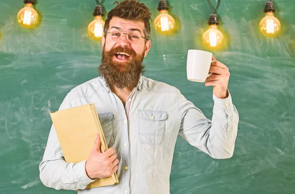 Hombre con barba en la cara feliz en el aula. Profesor en anteojos sostiene libro y taza de café o té. Concepto de pausa. Científico sostiene libro y taza de café, pizarra en el fondo, spac copia — Foto de Stock