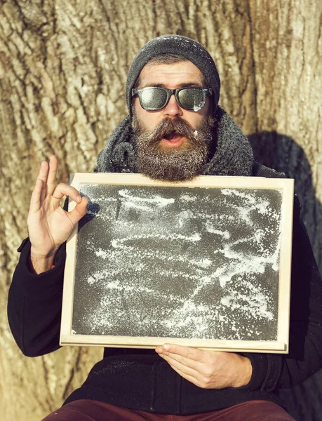 Upphetsad man, hipster, med skägg och mustasch i svarta solglasögon täckt med vit frost ger ok gest med svarta tavlan vinterdag på naturlig bakgrund, kopiera spac — Stockfoto