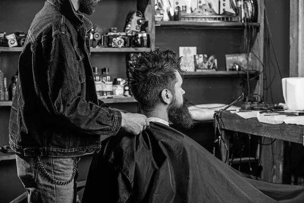 Man with beard and mustache sits in barbershop, beauty supplies on background. Barbershop concept. Man bearded client of hipster barbershop. Hipster with beard waits while barber cover neck with cape