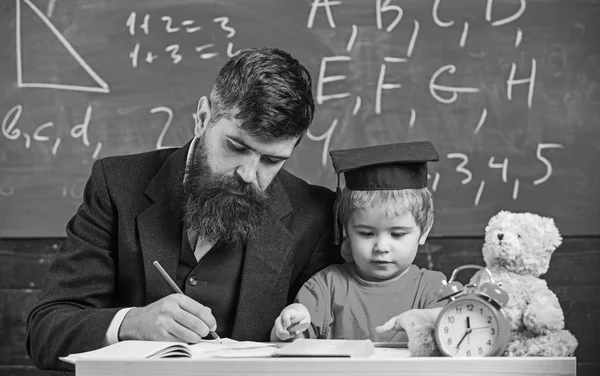 Busy kid studying at school. Teacher, father checking homework, helps to boy, son. Teacher in formal wear and pupil in mortarboard in classroom, chalkboard on background. Individual studying concept