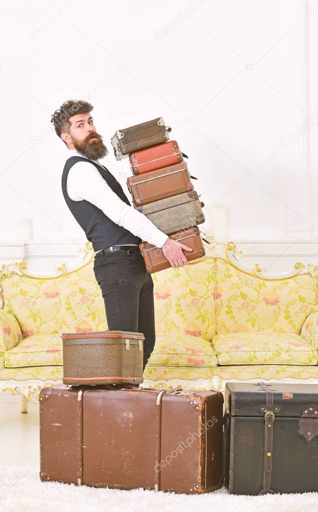Man with beard and mustache wearing classic suit delivers luggage, luxury white interior background. Butler and service concept. Macho, elegant porter on strict face carries pile of vintage suitcases