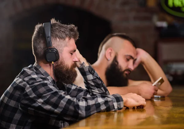 Friday relaxation in bar. Hipster bearded man spend leisure at bar counter. Order drinks at bar counter. Men with headphones and smartphone relaxing at bar. Avoid communication. Escape reality