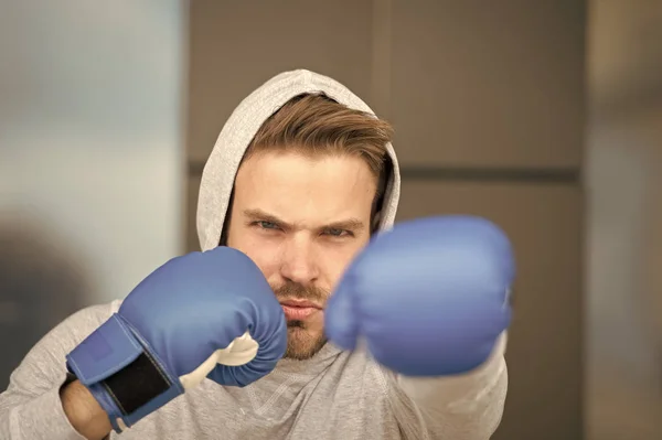 Boxing concept. Man athlete on concentrated face with sport gloves practicing boxing punch, urban background. Boxer with hood on head practices jab punch. Sportsman boxer training with boxing gloves