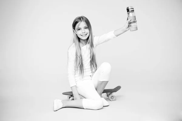 Niña cuidando la hidratación corporal. Ocio activo y equilibrio hídrico. Los niños activos y sanos beben agua. Mantenerse hidratado. Chica cara feliz sostiene con botella de agua mientras se sientan penny board fondo rosa — Foto de Stock