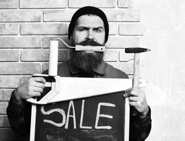 bearded foreman, long beard, brutal caucasian hipster, moustache, uniform, holding various building tools: saw, hammer, roller paint, board with inscription sale, serious face, brick wall backgroun