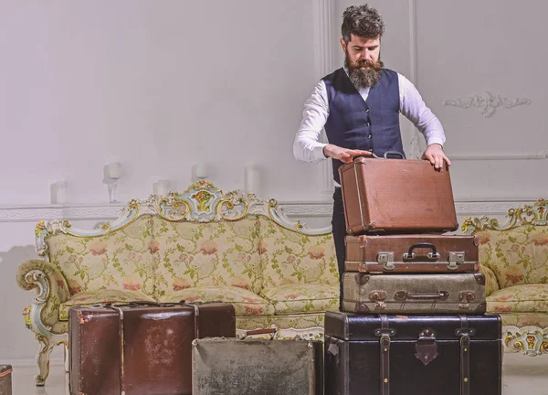 Hombre, viajero con barba y bigote equipaje de embalaje antes del viaje, fondo interior de lujo. Equipaje y concepto de viaje. Macho elegante en la cara reflexiva de pie cerca de pila de maleta vintage — Foto de Stock