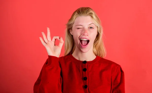 女孩眨眼快乐的脸, 而显示确定手势在红色的背景。女人对一切都满意。一切都很好或很好。好吧概念。好吧, 手势肢体语言。一切都会好起来的 — 图库照片