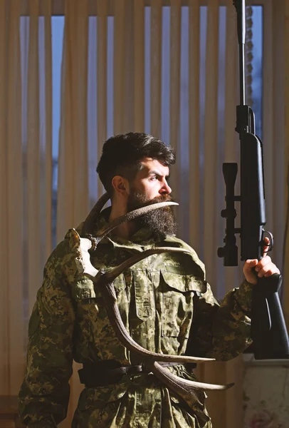 Wildhüter-Konzept. Mann mit Bart trägt Tarnkleidung, Vorhänge im Hintergrund. Jäger, brutaler Hipster mit Gewehr und Hirschhörnern. Macho mit strenger Miene im Jagdhaus — Stockfoto